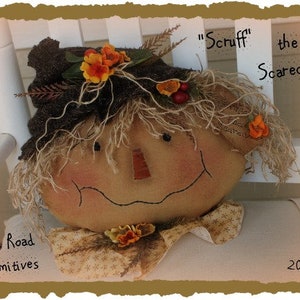 May include: A handmade scarecrow doll with a brown fabric hat, orange and yellow flower accents, and a friendly face. The doll is sitting on a white chair and is labeled "Scruff the Scarecrow".