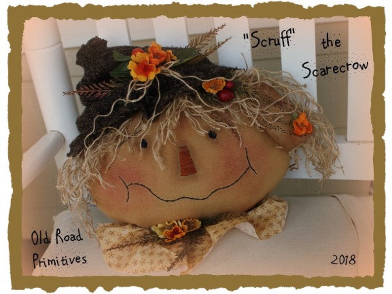 May include: A handmade scarecrow doll with a brown fabric hat, orange and yellow flower accents, and a friendly face. The doll is sitting on a white chair and is labeled "Scruff the Scarecrow".