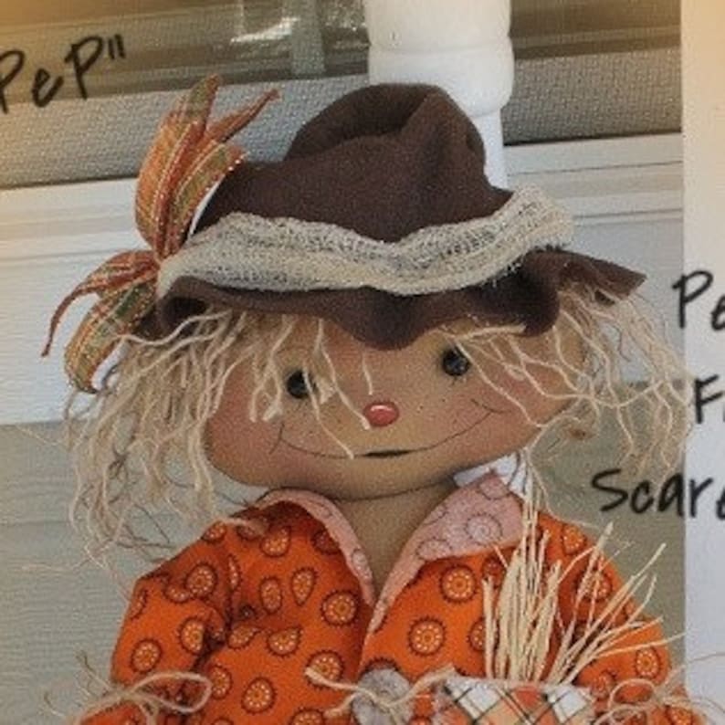 Puede incluir: Una mu&ntilde;eca de espantap&aacute;jaros hecha a mano con un sombrero de fieltro marr&oacute;n, una camisa con estampado naranja y blanco y cabello marr&oacute;n. La mu&ntilde;eca sostiene un manojo de paja.