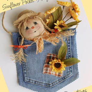 Op de afbeelding: Een denim zakhanger met een lachende pop met een strohoed en sjaal. De zak is versierd met zonnebloemen en een geruite patch. De tekst "Lil' Sunshine Girl Sunflower Pocket Hanger" en "Old Road Primitives" zijn zichtbaar.