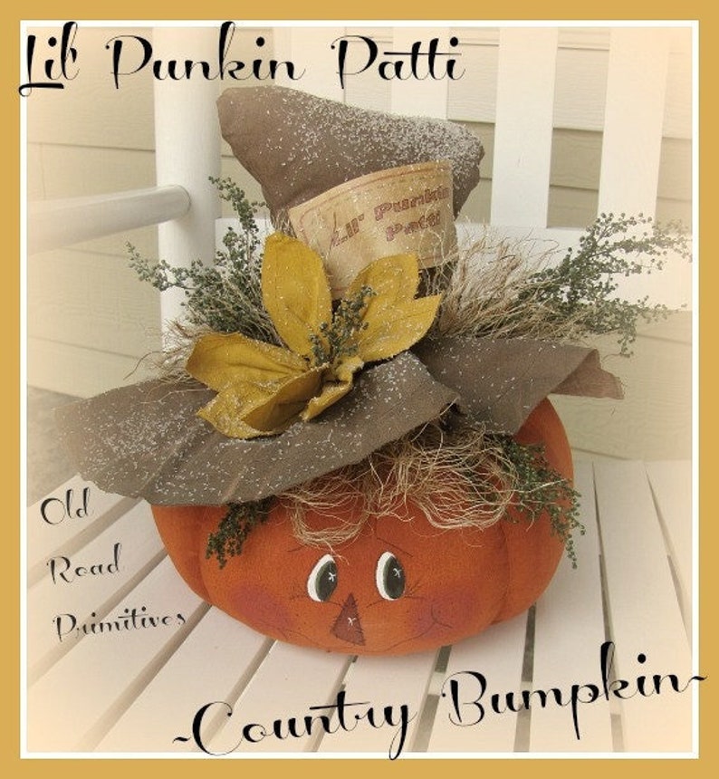 May include: A brown fabric pumpkin with a brown fabric hat and a yellow fabric flower on top. The pumpkin has a painted face with black eyes and a small mouth. The hat has a tag that says "Lil' Punkin Patti". The pumpkin is sitting on a white wooden bench. The text "Old Road Primitives - Country Bumpkin" is written below the pumpkin.
