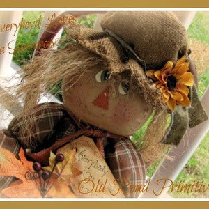 May include: A handmade rag doll scarecrow with a plaid shirt, brown hair, and a sunflower on its hat. The doll is holding a sign that says "Everybody Loves a Scarecrow".