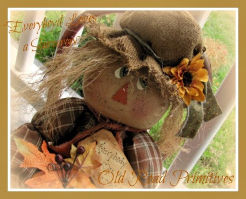 May include: A handmade rag doll scarecrow with a plaid shirt, brown hair, and a sunflower on its hat. The doll is holding a sign that says "Everybody Loves a Scarecrow".