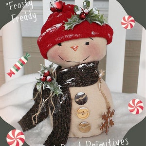 May include: A handmade snowman doll with a red hat, brown scarf, and holly accents. The doll has a button nose and is wearing a brown scarf. The doll is sitting on a white surface with a candy cane and snowflake.