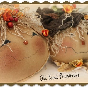 May include: Two handmade fabric scarecrows with painted faces and straw hair. The scarecrows are wearing fall-themed decorations, including flowers and berries. The scarecrows are sitting on a white surface with the text "Old Road Primitives" written below them.