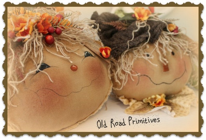 May include: Two handmade fabric scarecrows with painted faces and straw hair. The scarecrows are wearing fall-themed decorations, including flowers and berries. The scarecrows are sitting on a white surface with the text "Old Road Primitives" written below them.