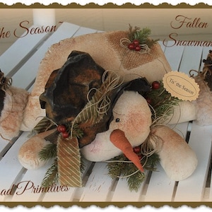 May include: A handmade fabric snowman doll with a brown hat, a carrot nose, and a green and brown garland. The snowman is lying on a white wooden surface. The text "Tis the Season" is written on the image.