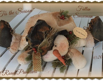May include: A handmade fabric snowman doll with a brown hat, a carrot nose, and a green and brown garland. The snowman is lying on a white wooden surface. The text "Tis the Season" is written on the image.