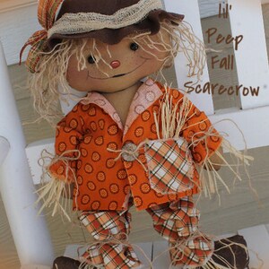 Puede incluir: Una mu&ntilde;eca de espantap&aacute;jaros de oto&ntilde;o hecha a mano con un sombrero de fieltro marr&oacute;n, pantalones de cuadros naranja y blanco y un sombrero de fieltro marr&oacute;n. La mu&ntilde;eca est&aacute; sentada en un banco de madera blanco. El texto "Lil' Peep Fall Scarecrow" est&aacute; escrito en el banco.