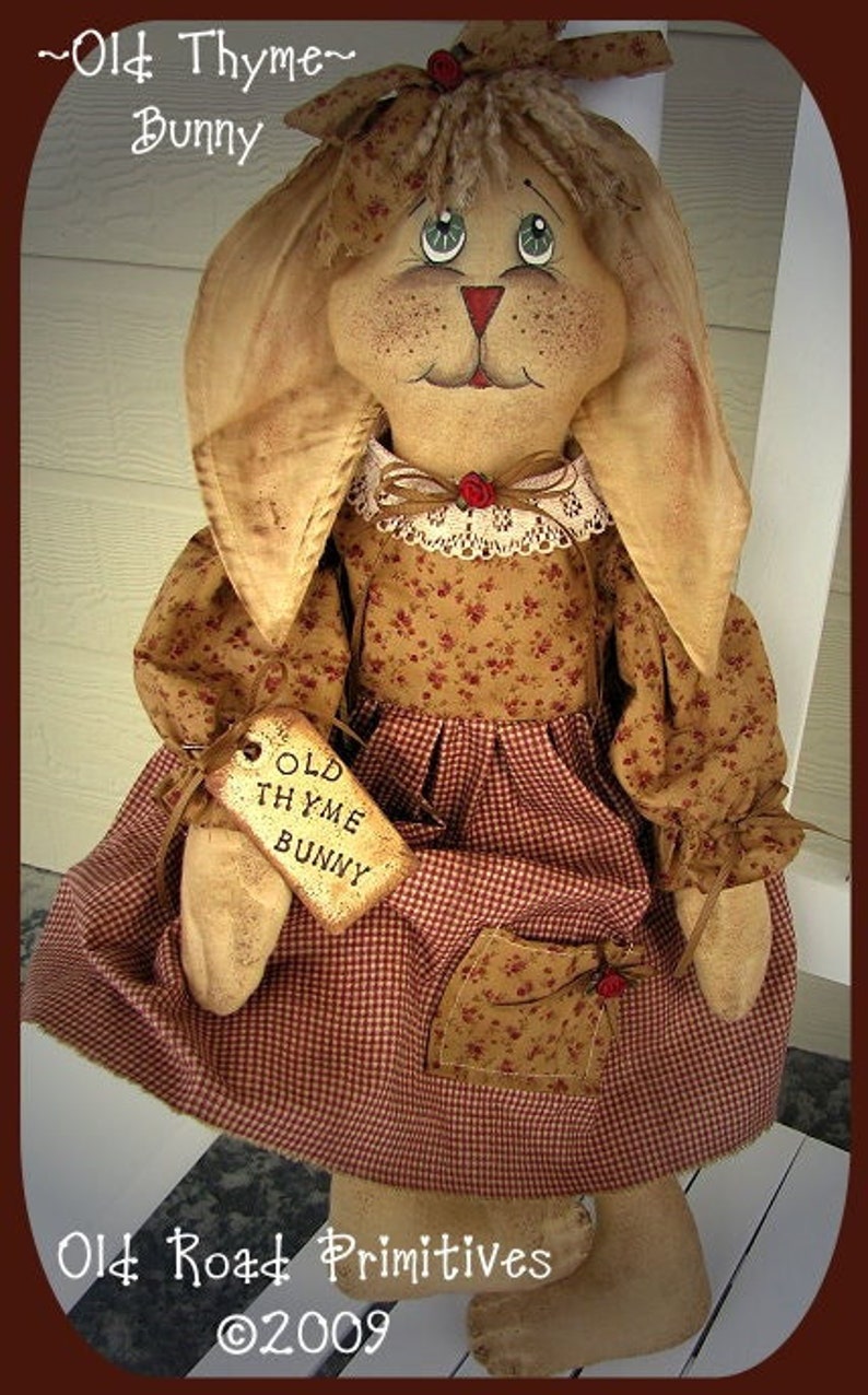 May include: A handmade fabric bunny doll with long ears, wearing a floral print dress and a red and white checkered skirt. The doll has a tag that reads "Old Thyme Bunny". The text "Old Road Primitives" and "&copy;2009" are also visible.