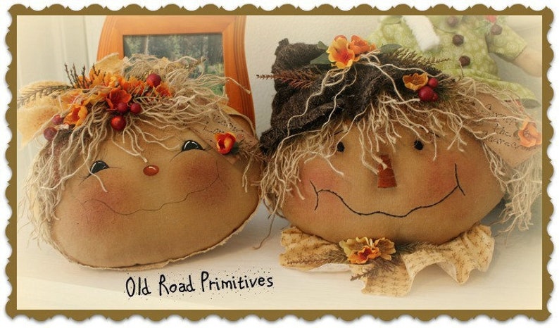 May include: Two handmade scarecrow figures with smiling faces, brown fabric bodies, and straw-like hair. The scarecrows are decorated with fall-themed accents, including orange and yellow flowers, and brown berries.