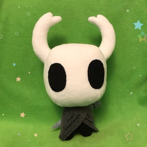Może przedstawiać: Biała pluszowa zabawka z czarnymi oczami i szarą peleryną. Zabawka jest postacią z gry wideo Hollow Knight.