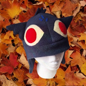 Puede incluir: Un gorro de forro polar azul con grandes ojos blancos y rojos y un par de orejas. El gorro está en una cabeza de maniquí blanca sobre un fondo de hojas de otoño.