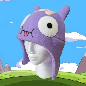May include: Purple fleece hat with a cartoon face, featuring a large white eye, a black pupil, a pink nose, and a pink tongue sticking out. The hat has a long flap that covers the ears and neck.