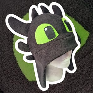 Puede incluir: Un gorro de forro polar negro y gris con forma de cabeza de dragón, con grandes ojos verdes y protuberancias en forma de orejas. El gorro tiene un contorno blanco y se muestra en una cabeza de maniquí.