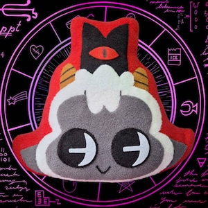 May include: A plush toy shaped like a gray and white creature with a red, black, and white hat. The creature has a single large red eye, two black eyes, and a small smile. The toy is against a black background with a pink circle pattern.