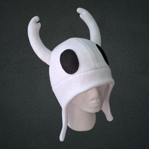 Puede incluir: Un gorro de forro polar blanco con dos cuernos grandes y curvados en la parte superior y dos parches negros ovalados prominentes en los laterales. El gorro tiene una corona redondeada y orejeras que caen hacia abajo, imitando una cabeza.