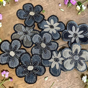 May include: A collection of nine fabric flower appliques in shades of charcoal gray, slate, and light gray. Each flower features five rounded petals with visible white and black decorative stitching, and a circular center with radiating thread details.