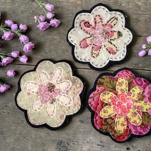 May include: Three fabric flower appliques with scalloped edges and layered petals in various floral patterns, including shades of pink, yellow, and cream, featuring visible hand-stitched details and dark fabric backings arranged with small purple flowers.