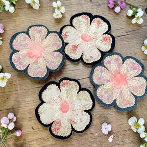 May include: Four flower-shaped fabric appliqués featuring a six-petal design with a central coral-colored circle. The petals display a mix of cream, pink, and purple floral patterns with visible stitching, all outlined by a dark navy blue border.