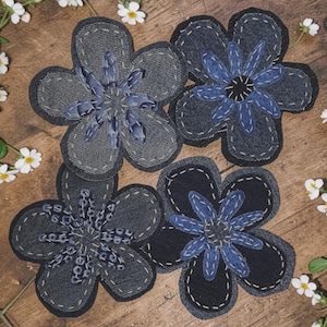 May include: Four handcrafted flower-shaped fabric patches made from dark blue and black denim. Each patch features five rounded petals with white decorative stitching along the edges and a layered center with intricate blue thread embroidery.