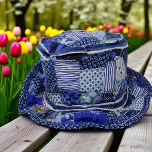 May include: A blue patchwork hat with a wide brim, featuring various patterns and shades of blue. The hat is made of quilted fabric and sits on a wooden bench, with a blurred background of colorful flowers.