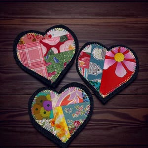 May include: Three heart-shaped fabric coasters with a patchwork design. Each coaster features a unique combination of patterned fabrics in various colors, including pink, red, green, and yellow, with a black border. The coasters are arranged on a dark wooden surface.