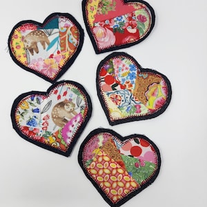 May include: Five handmade patchwork heart-shaped ornaments with a navy blue trim. Each heart is made of different colorful fabrics with floral and geometric patterns.