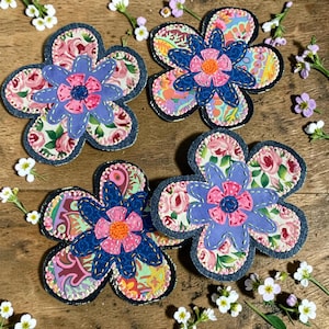 May include: Four fabric flower appliques featuring layered petals in various patterns including pink roses, paisley, and geometric prints. Each flower has a dark blue denim base, a light blue or dark blue top layer, and a small pink or orange center.