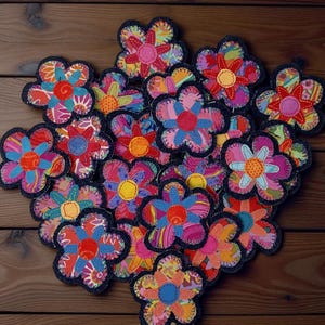 May include: A collection of colorful, handmade flower-shaped coasters. Each coaster features a unique patchwork design with vibrant hues of pink, blue, orange, and yellow, outlined with a dark blue border. The coasters are arranged on a wooden surface.