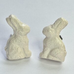 May include: Two white ceramic rabbit figurines. The rabbits are sitting upright with their ears perked up. They are both facing the right side of the image.