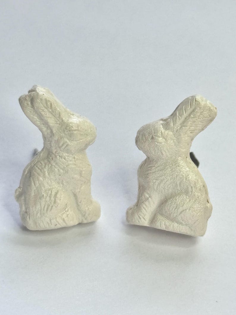 May include: Two white ceramic rabbit figurines. The rabbits are sitting upright with their ears perked up. They are both facing the right side of the image.