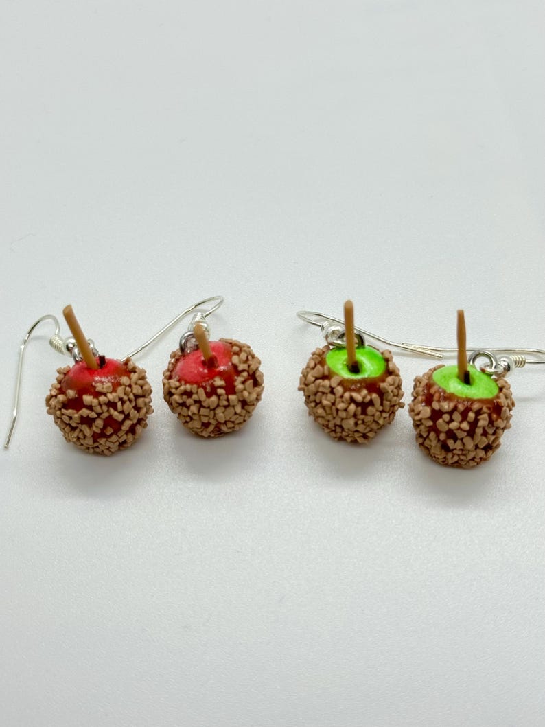 May include: A pair of handmade earrings featuring miniature candy apples. The earrings have a silver hook and are designed to look like caramel apples, with a red or green apple base covered in caramel and nuts. The earrings are on a white background.