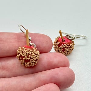 May include: A pair of miniature candy apple earrings. Each earring features a red apple base, coated in tan-colored candy pieces, and a small wooden stick. The earrings hang from silver-colored fish hook ear wires.