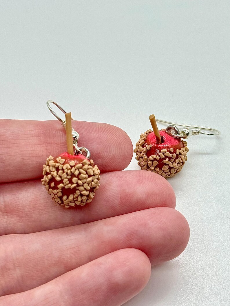 May include: A pair of miniature candy apple earrings. Each earring features a red apple base, coated in tan-colored candy pieces, and a small wooden stick. The earrings hang from silver-colored fish hook ear wires.