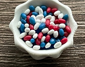 May include: A white bowl filled with red, white, and blue sprinkles. The sprinkles are in a variety of shapes and sizes.