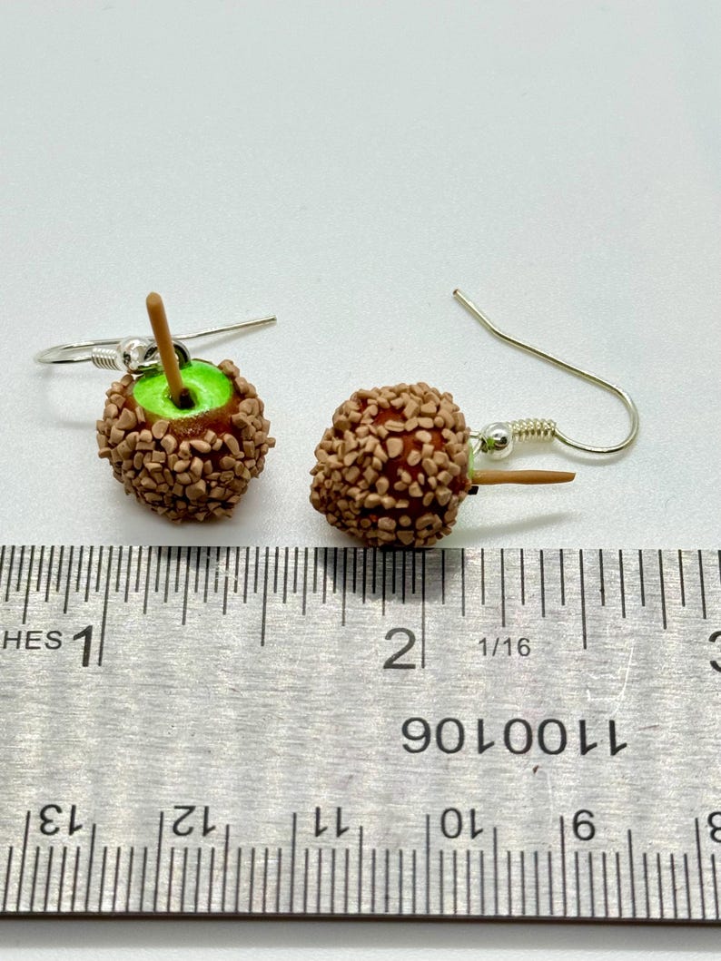 May include: A pair of handmade apple-shaped earrings. Each earring features a red apple covered in brown candy-like pieces, a green apple core, and a small wooden stick. The earrings hang from silver-colored fish hook ear wires.