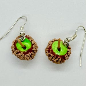 May include: A pair of dangle earrings featuring miniature caramel apples. Each earring has a bright green apple base, coated in a layer of caramel and chopped nuts, with a small wooden stick and silver-colored ear wire.