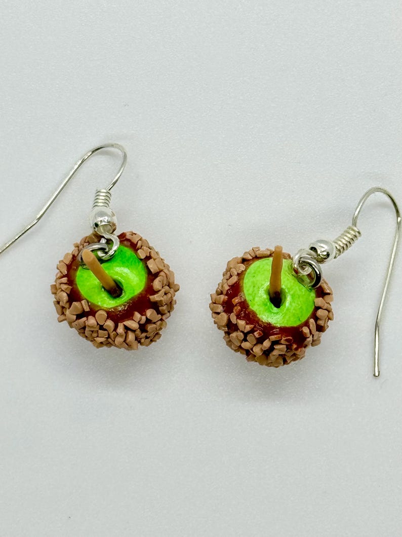 May include: A pair of dangle earrings featuring miniature caramel apples. Each earring has a bright green apple base, coated in a layer of caramel and chopped nuts, with a small wooden stick and silver-colored ear wire.