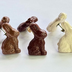 May include: Three pairs of bunny rabbit stud earrings. One pair is brown, one pair is white, and one pair is a dark brown color.