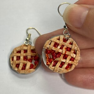 May include: A pair of cherry pie earrings. The earrings are made of polymer clay and are shaped like a cherry pie. The pie crust is a light brown color and the cherries are a bright red color. The earrings are hanging from silver hooks.