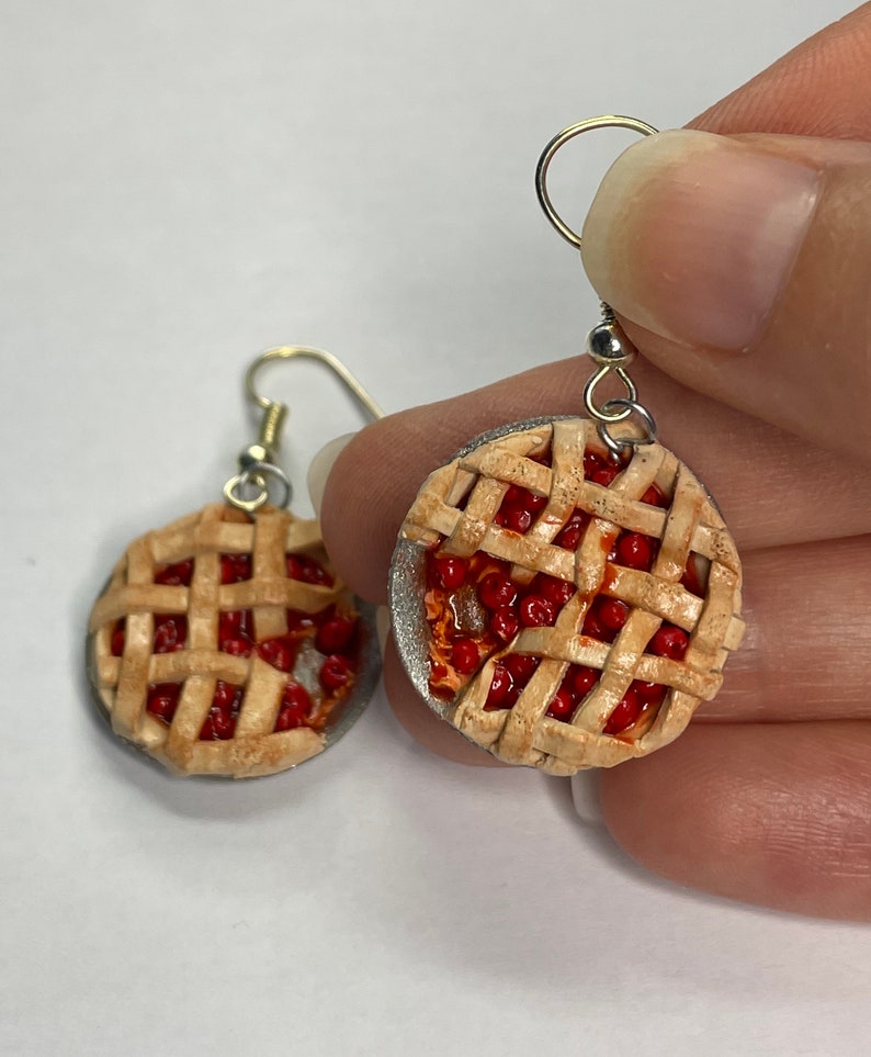 May include: A pair of cherry pie earrings. The earrings are made of polymer clay and are shaped like a cherry pie. The pie crust is a light brown color and the cherries are a bright red color. The earrings are hanging from silver hooks.