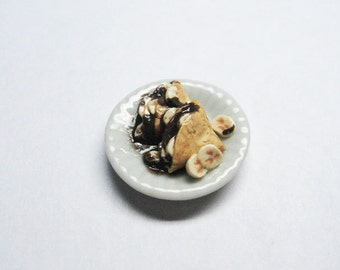 Può includere: Un piatto bianco con una porzione di crêpes in miniatura ricoperte di salsa al cioccolato e fette di banana.