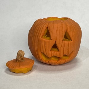 Pu&ograve; includere: Una zucca arancione intagliata con un ghigno dentato e un coperchio rimovibile. La zucca &egrave; realizzata in un materiale strutturato che ricorda il legno.
