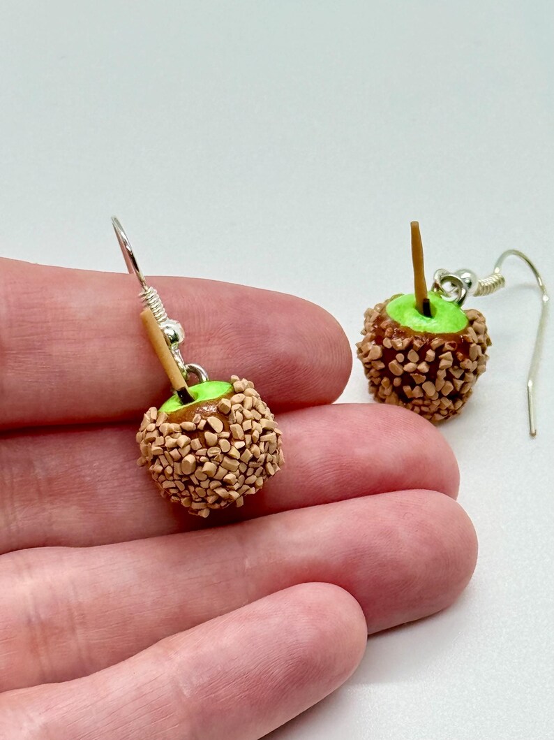 May include: A pair of handmade apple-shaped earrings. Each earring features a green apple base covered in small, brown, candy-like pieces, with a small wooden stick. The earrings hang from silver-colored hooks.