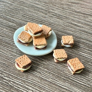 以下が含まれることがあります： グラハムクラッカー、チョコレート、マシュマロで作ったミニスモアが載ったプレート。スモアは白いプレートに盛り付けられ、茶色の木製の背景があります。