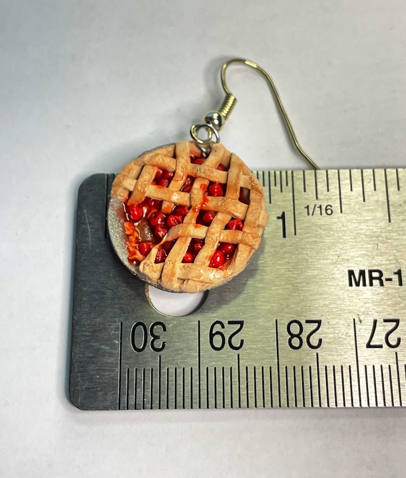 May include: A pair of cherry pie earrings, each with a red and brown pie crust and a red cherry filling. The earrings are about 1 inch in diameter and are shown next to a ruler.