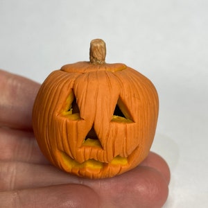 Pu&ograve; includere: Una piccola zucca arancione e gialla con un viso intagliato. La zucca &egrave; fatta di argilla e ha una superficie testurizzata.