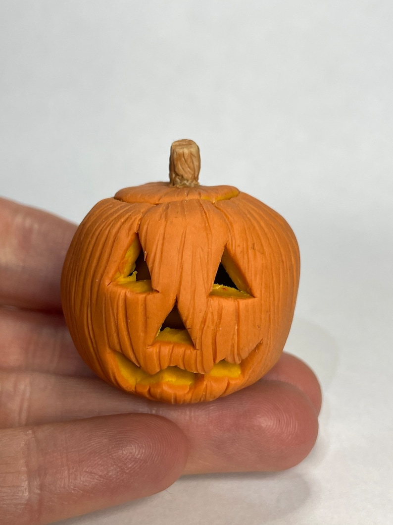 Pu&ograve; includere: Una piccola zucca arancione e gialla con un viso intagliato. La zucca &egrave; fatta di argilla e ha una superficie testurizzata.