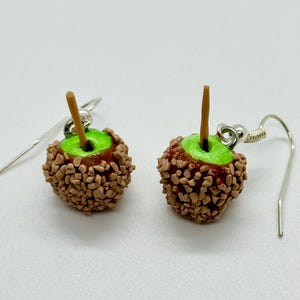 May include: A pair of handmade caramel apple earrings. Each earring features a miniature green apple base, coated in a brown candy shell with a wooden stick. The earrings have silver-colored ear wires.